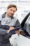 Portrait of confident mechanic holding clipboard while leaning on car's window in workshop