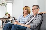 Parents watching TV on sofa with children using laptop in background