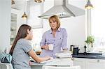 Mother having coffee while looking at daughter studying in kitchen