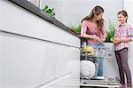 Happy mother and daughter placing glasses in dishwasher at kitchen