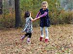 Two little girls hold hands and swing in circles.  They are playing outdoor games in the Fall leaves.