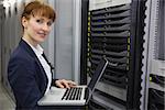 Pretty technician using laptop while working on server smiling at camera in large data center