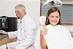 Smiling little girl showing thumbs up in dentists chair at the dental clinic