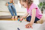 Little girl drawing in the living room with mother sitting behind at home