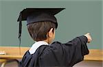Cute pupil in graduation robe pointing in classroom at the elementary school