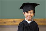 Cute pupil in graduation robe smiling at camera in classroom at the elementary school