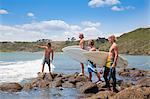 Four young male surfer friends pointing to sea from rocks
