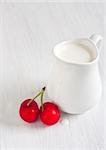 Cream in little white jug on white wooden table with red cherry. High key.