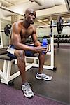 Full length of muscular man with energy drink sitting in gym