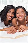 Mother and daughter reading book together on bed at home in bedroom