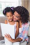 Pretty mother sitting on couch with cute daughter using tablet together at home in the living room