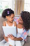 Pretty mother sitting on couch with cute daughter using tablet together at home in the living room