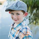 A young boy wearing a checked shirt and cloth cap with a large brim standing on a riverbank.