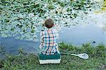 Back view of a young boy sitting on his baitbox, fishing in the river.