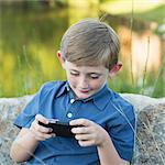 A young boy outdoors sitting leaning against a rock, using a handheld electronic game.