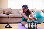 Young woman exercising on sitting room floor whilst looking at digital tablet