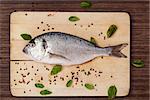 Sea bream on wooden chopping board with colorful peppercorns and basil leaves on brown background. Culinary seafood concept in natural brown.