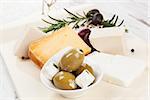 Different cheese, black olives, rosemary and colorful peppercorns on kitchen board on white wooden background. Culinary eating background.