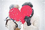 Attractive young couple in warm clothes holding red heart against snow falling