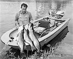 1970s MAN IN SMALL MOTORBOAT AT EDGE OF LAKE HOLDING UP CATCH OF THREE LARGE MUSKIES