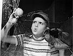 1950s BOY IN TEE-SHIRT & CAP REMOVING BASEBALL FROM BROKEN WINDOW