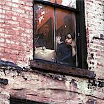 1960s 1970s SINGLE SAD LITTLE GIRL SITTING IN WINDOW OF WORN URBAN BRICK BUILDING