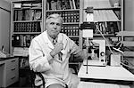 1980s SENIOR MALE DOCTOR SITTING AT DESK BEHIND MICROSCOPE GLASSES REMOVED TURNED LOOKING AT CAMERA