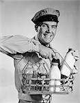 1950s 1960s SMILING MILKMAN IN UNIFORM LOOKING AT CAMERA CARRYING TWO FULL AND TWO EMPTY BOTTLES OF MILK IN METAL CARRIER