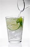 Clear water pouring from jug into drinking glass with lime and ice cubes