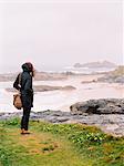 A woman lookng over the rocks and the shoreline on a windy day by the sea.