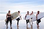 Group of male and female surfer friends walking away from sea with surf boards