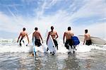 Rear view of a group of male and female surfer friends wading into sea with surf boards