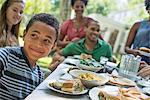 A family gathering, men, women and children around a table in a garden in summer. A boy smiling in the foreground.