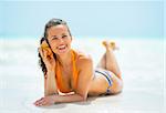 Happy young woman laying on sea shore and listening sound of sea in shell