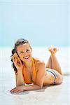 Smiling young woman laying on sea shore and listening sound of sea in shell