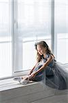 Little ballerina in gray dress puts on ballet shoes pointe front of the window.