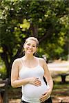 Portrait of happy pregnant woman touching belly and standing at camera smiling in city park
