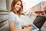 Smiling pretty redhead typing on laptop in the street