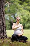 Portrait of white beautiful pregnant woman doing yoga exercise in park near tree, touching belly and heart
