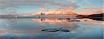 Panoramic view across the calm water of Jokulsarlon glacial lagoon towards snow-capped mountains and icebergs bathed in late afternoon light in winter, at the head of the Breidamerkurjokull Glacier on the edge of the Vatnajokull National Park, South Iceland, Iceland, Polar Regions