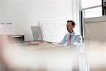 A man seated at an office desk using a computer. A wall chart with post it adhesive notes.