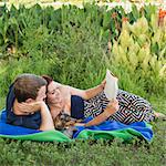 A couple, man and woman sitting on a picnic rug reading a book with their small dog between them.