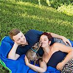 A couple sitting on a picnic rug. A small dog licking the face of a woman.