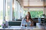 A young woman seated at a table in a diner, wearing a straw hat Using a digital tablet.