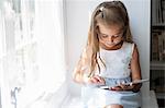 A young girl sitting at a window seat, using a digital tablet.