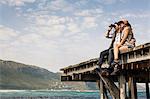 Young couple sitting on edge of old pier looking through binoculars, Cape Town, Western Cape, South Africa