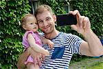 Mid adult man with baby daughter taking selfie on smartphone  in garden