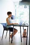 Mid adult mother typing on laptop with toddler daughter under the table