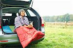 Woman sitting at rear of car with legs tucked in sleeping bag