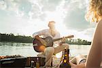 Young man sitting by lake playing guitar